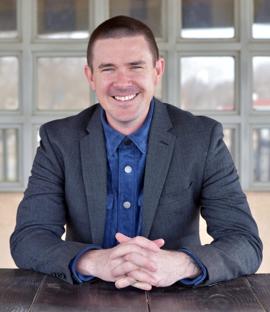 Full color vertical photo of Levi Monagle at a table with fingers interlaced, smiling into the camera
